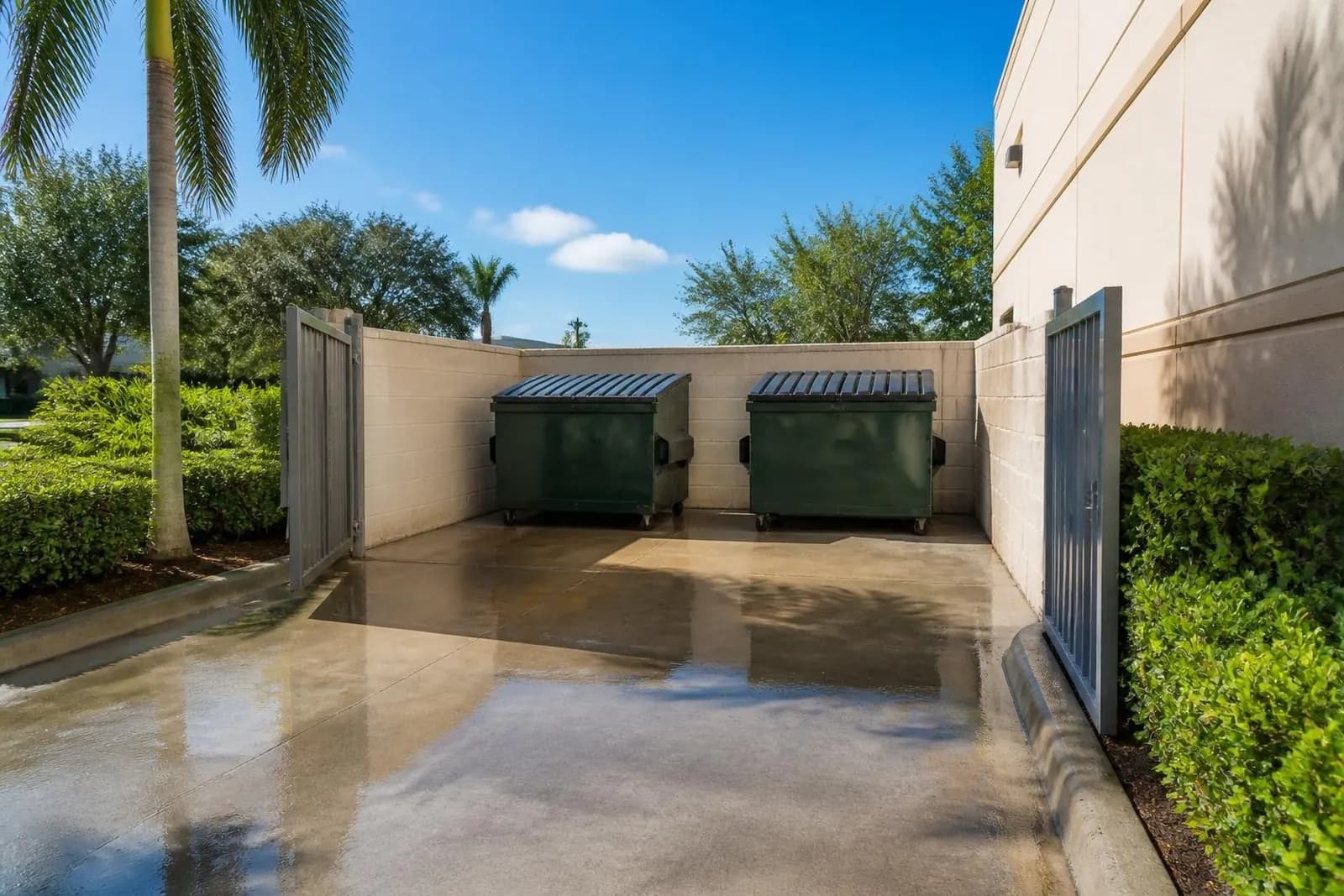 A freshly pressure-washed concrete dumpster pad with closed dumpsters inside a block enclosure behind a commercial building